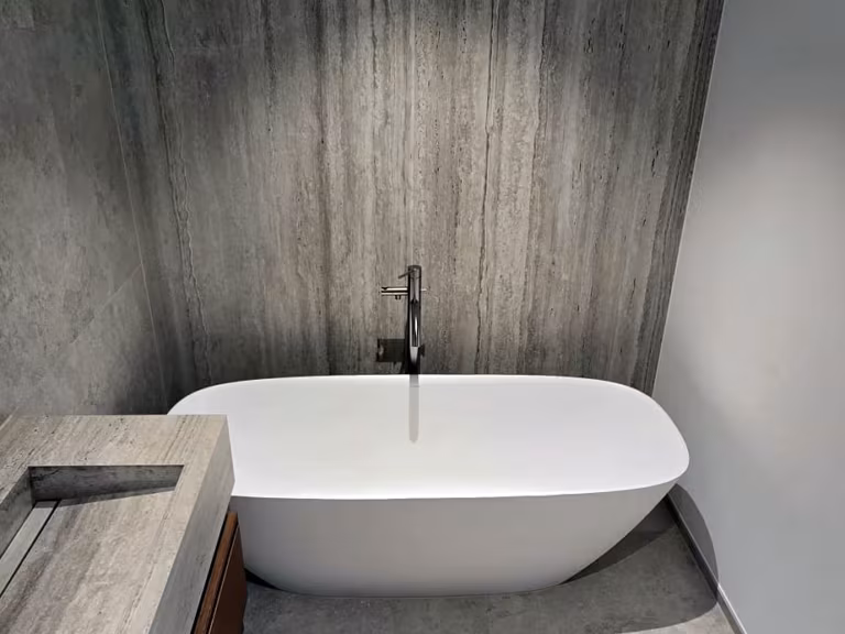 Freistehende Badewanne mit Standarmatur vor raumhohen Wandplatten in Travertinoptik